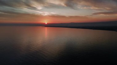 Gün batımı, kıyı şeridi üzerinde canlı turuncu ve mor renkler oluştururken, su üzerinde yansımalar parıldıyor. Narin bir sis tabakası kıyıya yakın duruyor. Huzurlu sahneyi güzelleştiriyor.