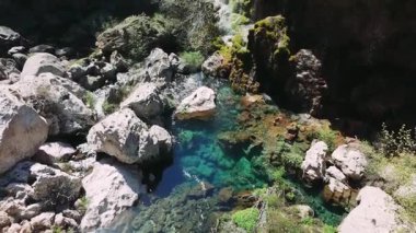 Şelale gibi kristal su, yosunlu taşların üzerine yuvarlanarak güneş ışığıyla aydınlanan kayalık kanyon manzarasına gömülü yeşil zümrüt havuzu oluşturur.