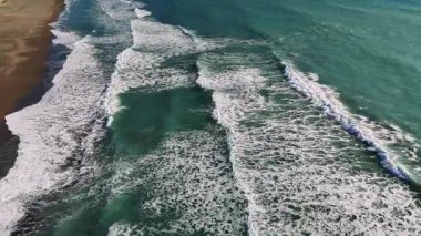 Turkuaz okyanus dalgaları ritmik bir şekilde yuvarlanıyor ve kumlu sahile çarpıyor. Havadan bakıldığında büyüleyici doğal deniz manzarası dinamikleri ortaya çıkıyor.