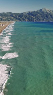 Turkuaz dalgalar, altın kumlu kıyı şeridine hafifçe çarpıyor. Parlak güneş ışığı altında, el değmemiş Akdeniz suları altında serin kıyı manzarasını yansıtıyor.