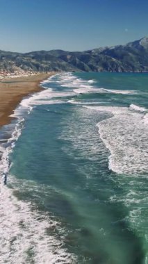 Turkuaz dalgalar yavaşça kumlu kıyı şeridine çarpıyor, parlak yaz gökyüzü altında ihtişamlı arka planda yükselen manzaralı dağ sporları, huzurlu Akdeniz kıyı manzarasını yakalıyor