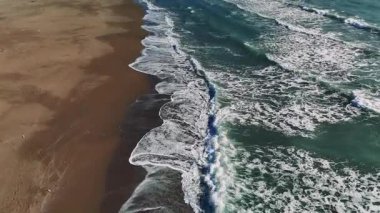 Turkuaz okyanus suyu beyaz köpük oluştururken, yumuşak dalgalar parlak güneş ışığı altında el değmemiş kumlu kıyı şeridi üzerinde yuvarlanırken, durgun deniz manzarası sunar.