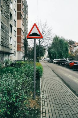 İkamet binalarıyla çevrili ve park halindeki arabalar ve ağaçlarla çevrili bir şehir caddesinde hız tümseği için uyarı işareti var..