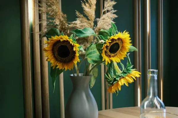Sarı ayçiçekleri ve otlar bir vazoyu ahşap bir masadan doldurur. Yeşil bir duvarın yanında berrak bir şişe duruyor..