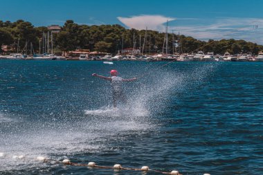 Genç bir çocuk, güneşli bir yaz öğleden sonrasında parlak mavi göl suyunda süzülerek bir wakeboard üzerinde ustaca manevra yapar.. 