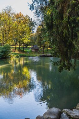 Ziyaretçiler, yerel bir tarih müzesini, sazdan yapılmış ahşap bir evi ve ağaçlarla çevrili gölet yüzeyinde parıldayan yansımaları sergileyerek huzurlu bir parkta geziniyorlar..
