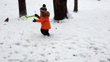 Sakin bir kış parkında, parlak turuncu paltolu genç bir çocuk renkli oyuncak küreğini kullanarak neşeyle karları havaya fırlatıyor. Kahkahalar çalarken serin havayı doldurur.