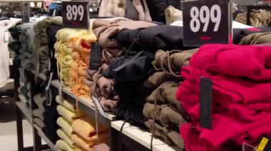 Raflar düzgün katlanmış kıyafetlerle dolu ve canlı renkler dikkat çekiyor. Her parça açık bir fiyat etiketi içeriyor, müşterileri çeşitli tarz ve renkleri keşfetmeye davet ediyor.