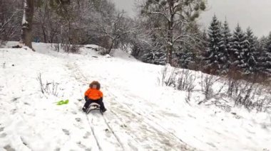 Heyecan dolu küçük bir çocuk, parktaki karlı tepeden kayarak serin bir kış gününün tadını çıkarıyor. Onun parlak turuncu kıyafeti soğuk manzaraya karşı duruyor, neşeli bir an yaratıyor..