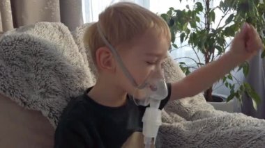 Küçük bir çocuk solunum tedavisi için solunum maskesi takan yumuşak bir koltuğa rahatça oturur. Güneş ışığı içeri süzülür ve odada sıcak, sakin bir atmosfer yaratır..