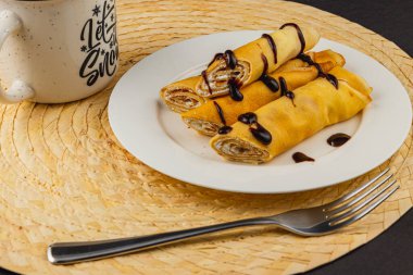 İçi iyilikle dolu sıcak, rulo krepler beyaz bir tabakta zarifçe otururlar. Zengin bir çikolata serpintisi davetkar bir dokunuş ekler, yakınındaki rahat bir kupayla tamamlanır, enfes bir aperatif için mükemmeldir..