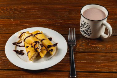 Delicate sweet crepes are served on a white plate, generously drizzled with rich chocolate syrup. A cozy cup of hot cocoa sits nearby, inviting warmth on a wooden table. clipart