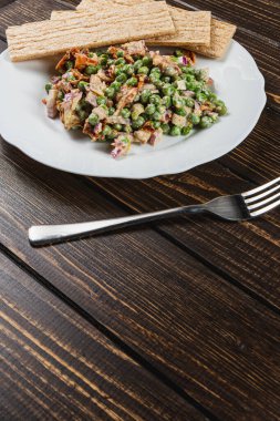 Lezzetli bir kase yeşil bezelye salatası, mor soğan ve soğan karışımı beyaz bir tabakta duruyor, yanında da tahıllı ekmek var, hepsi güzel bir şekilde sade ahşap bir masa üzerine yerleştirilmiş..