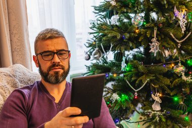 Genç bir adam rahat bir sandalyede rahatlar, e-kitap okumaya dalmış, festival ışıkları yakındaki Noel ağacında parıldarken, sıcak ve davetkar bir atmosfer yaratır..