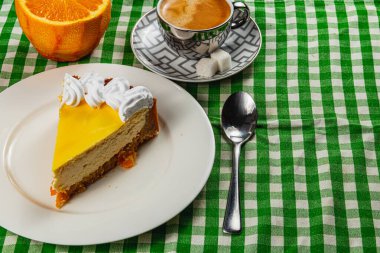 Bir dilim ev yapımı portakallı cheesecake, dumanı tüten bir fincan espresso ile tamamlanmış bir tabağın üzerine davetkâr bir şekilde oturur. Taze portakal yarımları sıcak sahneye canlı bir dokunuş ekler..