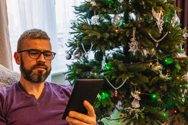 Genç bir adam e-kitabına dalmış bir sandalyede rahatça oturuyor. Yakınlarda, güzel dekore edilmiş bir Noel ağacı ışık ve süslemelerle parıldıyor ve rahat bir tatil atmosferi yaratıyor..