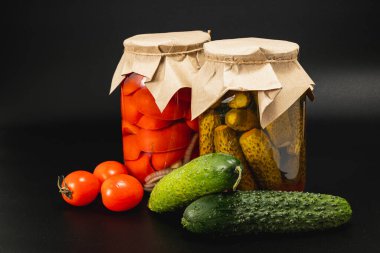 Two jars filled with homemade pickled cucumbers and vibrant tomatoes are surrounded by fresh vegetables. The jars feature decorative paper lids, enhancing their rustic charm. clipart