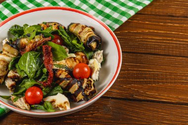 Izgara sebzeler, taze ıspanak ve sulu vişne domatesleriyle dolu canlı bir salatayla dolu bir kase, yakınlardaki yeşil kareli kumaşlı ahşap bir masaya kurulmuş..