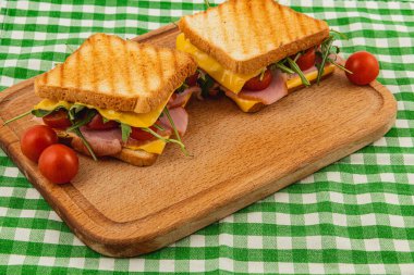 Eritilmiş peynir, jambon, ıspanak, roka ve taze domatesli iki ızgara sandviç ahşap bir tahtanın üzerinde oturur. Bu ev yapımı yemek güneşli bir günde öğle yemeği veya brunch için mükemmeldir..