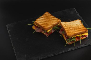 İki ızgara peynirli sandviç. Katmanlı jambon, taze ıspanak, roka ve sulu domatesler siyah bir taş tabakta oturuyorlar. Atıştırmalık ya da hafif öğle yemeği için mükemmel..