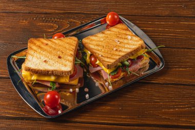 A platter features two grilled sandwiches stuffed with cheese, ham, fresh spinach, arugula, and cherry tomatoes, highlighting a wholesome snack perfect for lunch or brunch. clipart