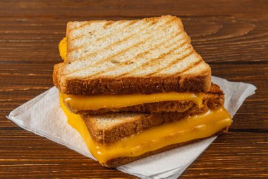 Lezzetli bir ızgara peynirli sandviç, kırsal ahşap bir masanın üstündeki kağıt peçetenin üzerinde oturur. Günün her saatinde keyif almaya davet eder. Kahvaltı ya da aperatif için mükemmeldir..