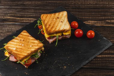 İki ızgara sandviç, içi jambon, duble peynir, domates ve yeşillik dolu siyah bir tabakta servis edilir. Öğle yemeği ya da kahvaltı için cazip bir seçim olur. Taze vişneli domatesler yemeğe eşlik eder..