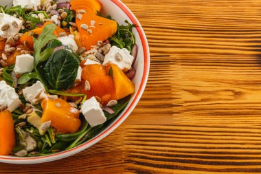 A vibrant salad showcases fresh ingredients like persimmon, feta cheese, and sunflower seeds on a wooden table. clipart