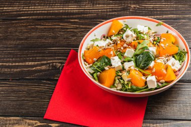 İçinde hurma, ıspanak, roka, tavuk filetosu, feta peyniri, mor soğan ve ahşap bir masada servis edilen ayçekirdeği bulunan canlı bir salata..