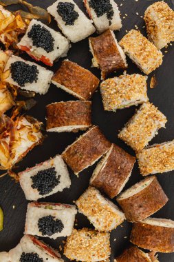 Çeşitli suşi ruloları, havyar ve susam gibi malzemelerin sergilendiği siyah bir yüzeyde sanatsal olarak düzenlenir. Ekranda, çevredeki malzemelerin turuncu ve kahverengi aksanları yer alıyor..