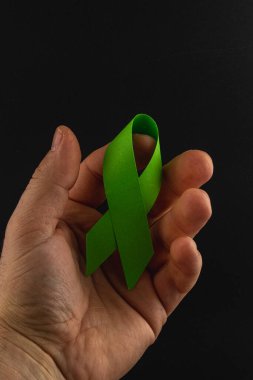 Bir el, kanserle mücadeleyi sembolize eden yeşil bir farkındalık şeridi tutuyor. Bu, umut, destek ve sağlık hizmetleri farkındalığı ve önleme çabalarına duyulan ihtiyacı temsil etmektedir..