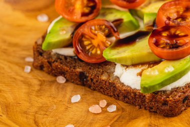 Olgun avokado ve vişneli domates dilimleri bir parça siyah ekmek üzerinde ustalıkla düzenlenir. Kırsal ahşap bir tahtaya serpiştirilmiş tuz taneleri taze malzemeler sergiliyor..