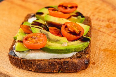 Taze hazırlanmış avokado tostu, olgun avokado dilimleri, vişneli domatesler ve balzamik jöleli bir çiseleme içeren ahşap bir tahtaya kaplanmış. Kahvaltı ya da hafif atıştırmalık için mükemmel..