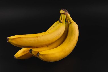 Olgun sarı muz kümesi, parlak renklerini ve pürüzsüz dokularını vurgulayan koyu bir arka planda bulunur. Düzenleme meyvelerin doğal kıvrımlarını ve çekici şekillerini gösteriyor..