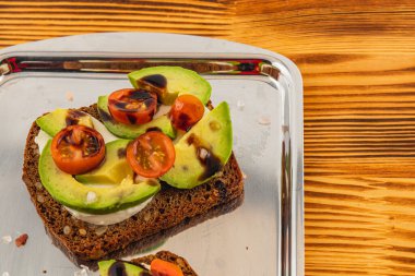 Lezzetli bir açık yüzlü sandviç siyah ekmeğin üzerinde taze avokado dilimleri ve kiraz domatesleri barındırır. Sunum davetkar, ahşap bir zemine karşı parlak gümüş bir tepsiye kurulmuş..