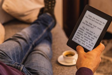 Kanepede oturup e-kitap okuyan biri. Bir fincan kahve yakınlarda oturuyor, kitap okuma haftasında hikayelerden zevk almak için mükemmel bir ortam yaratıyor..