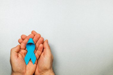 Bir hasta, her iki elinde de gri bir arkaplana karşı mavi kurdele tutarak, prostat kanseri tespiti ve sağlık hizmetlerindeki önleme çabalarına verilen bilinci ve desteği temsil ediyor..