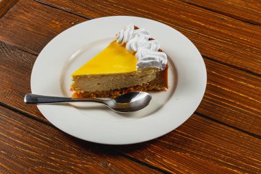 Lezzetli bir dilim cheesecake yanında kaşıkla beyaz bir tabakta servis edilir. Sarı jöle canlı bir dokunuş eklerken, krem şanti çekiciliğini artırıyor..