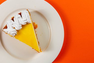 Süslü bir turta dilimi beyaz bir tabakta duruyor, pürüzsüz sarı bir dolgu sergiliyor ve üzerinde parlak turuncu bir zemin üzerine krema dökülüyor..