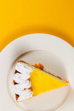 Canlı bir dilim limonlu turta, üzerinde krem şanti olan beyaz bir tabağın üzerinde duruyor. Parlak sarı arka plan, güneşli tatlının neşeli görünümünü güzelleştirir..