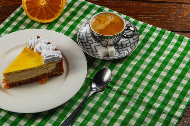 Krem şanti ile süslenmiş canlı bir dilim turunçgil peyniri beyaz bir tabakta, dumanı tüten bir fincan espressonun yanında duruyor. Masada yeşil kareli kumaş ve bir dilim portakal var..