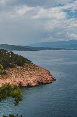 Engebeli bir kıyı şeridinin kıyısında, hafif dalgalar, günışığı altındaki kayalara çarpar. Bereketli yeşillik bulutlu bir gökyüzünün altındaki sakin suları çerçeveler ve okyanus tarafından barışçıl bir kaçış yaratır..