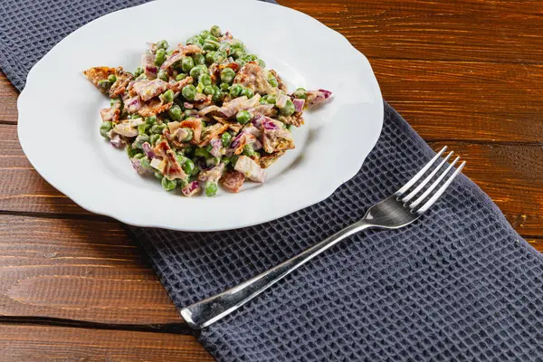 Beyaz bir tabakta taze bezelye, sebze ve çıtır çıtır malzemelerden oluşan canlı bir karışım. Yemek ahşap bir masada, koyu mavi bir peçete ve parlak bir çatalla hazırlandı..