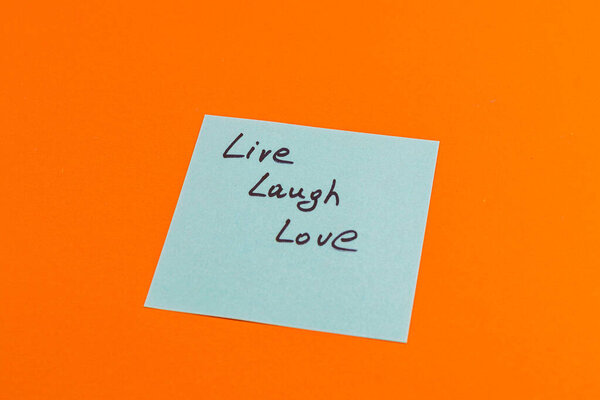 A light blue sticky note bears the handwritten words 'Live Laugh Love' in bold black letters, resting on a bright orange surface, conveying positivity and motivation.