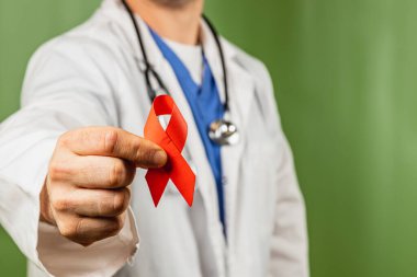 Bir sağlık çalışanı, yeşil zemine karşı HIV AIDS farkındalığını sembolize eden kırmızı bir kurdele tutuyor. Bu kişi steteskop takıyor ve tıp mesleğini vurguluyor..