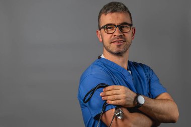 Mavi önlüklü bir adam, koluna steteskop sarılı bir şekilde duruyor. Klinik bir ortamda profesyonellik ve hasta bakımına olan bağlılığını gösteriyor..