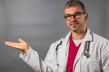 Beyaz önlüklü ve gözlüklü bir doktor uzanmış elleriyle el kol hareketi yapıyor. Ayarlar, profesyonelliğin ve yaklaşılabilirliğin vurgulandığı klinik bir ortam izlenimi veriyor.