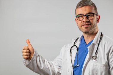 Neşeli bir doktor beyaz önlük ve mavi önlük giyerken başparmağını kaldırır. Klinik ortamda, sağlık hizmetlerinde olumlu bir atmosfer vurgulanıyor..