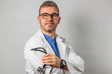 Erkek bir doktor steteskop tutarken kendinden emin bir şekilde gülümser. Beyaz önlük ve mavi önlük giyerek, klinik bir ortamda profesyonelliği ve ulaşılabilirliği aktarıyor..