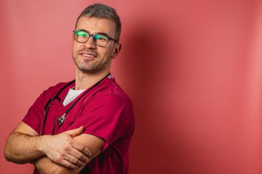 Kırmızı önlük ve gözlük takan bir adam kollarını kavuşturup güvenle gülümsüyor. Steteskopu boynunda asılı ve parlak pembe bir arka plana karşı, profesyonellik saçıyor..